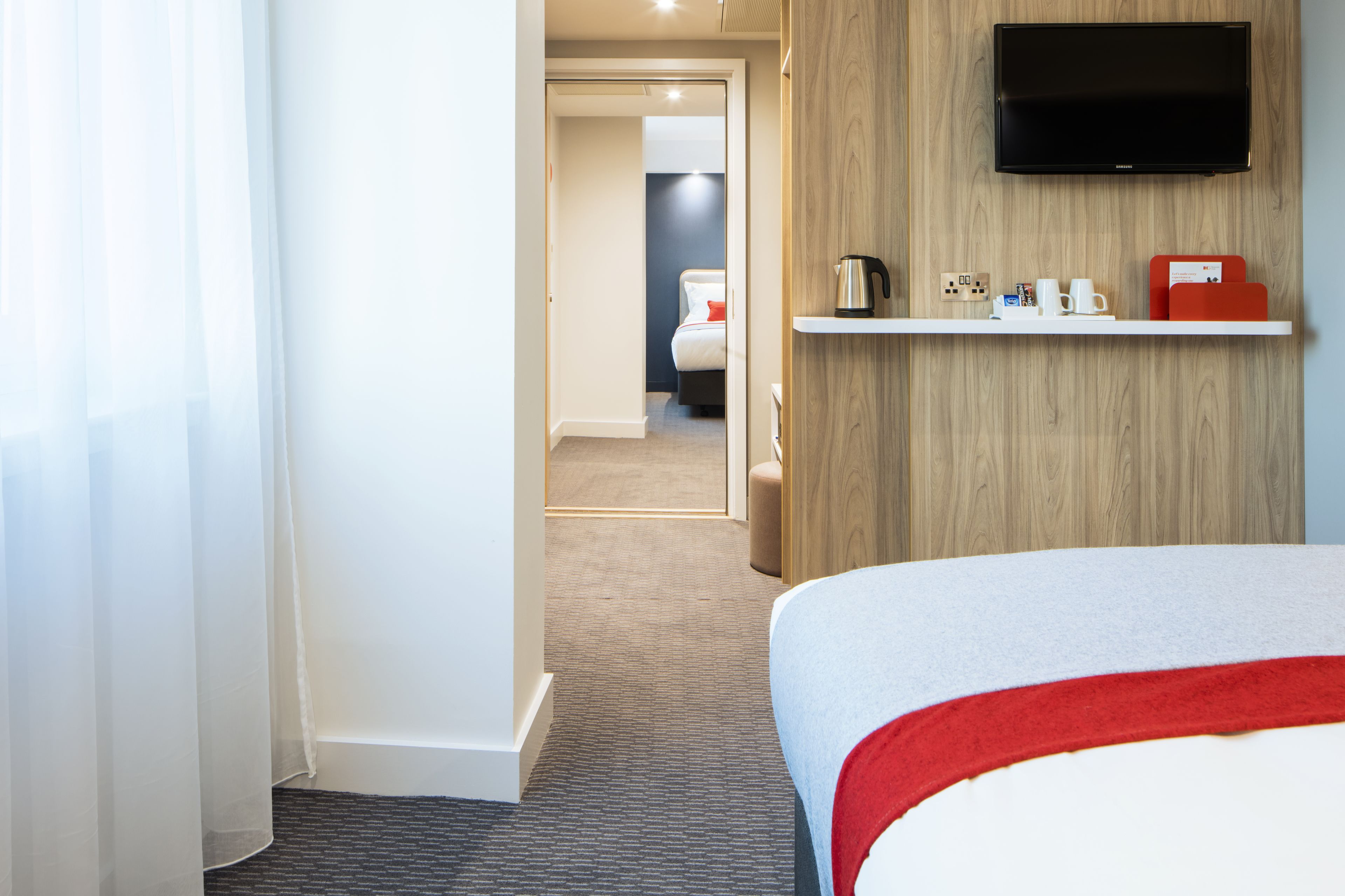 A hotel room with a bed, a television mounted on the wall, a shelf with a coffee maker, cups, and a red box.