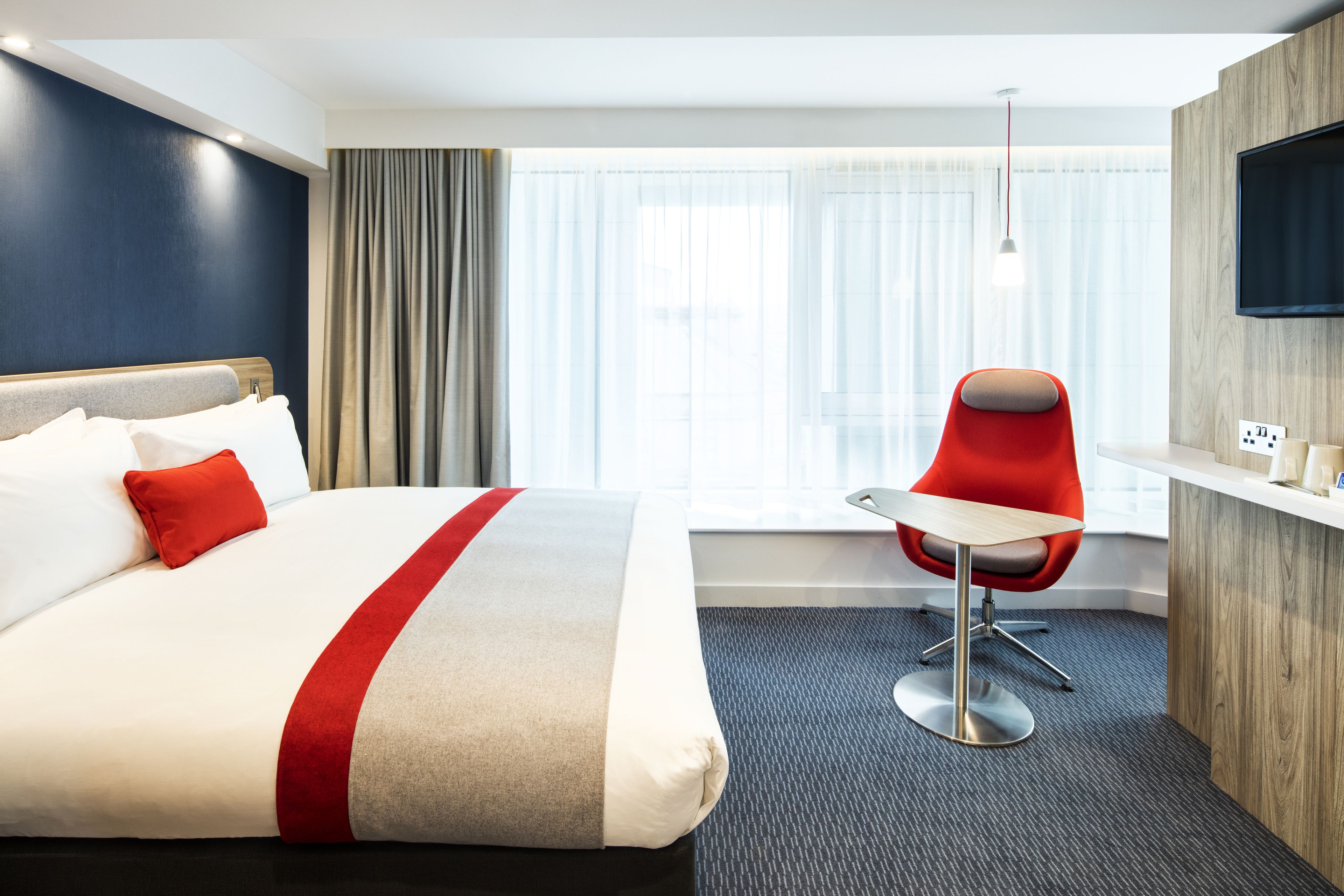 A hotel room with a large bed, a red chair, a small table, and a TV mounted on the wall.