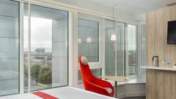 Una habitación de hotel moderna con ventanas amplias, una silla roja, una mesita pequeña y un televisor montado en la pared.