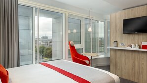 Ein modernes Hotelzimmer mit einem großen Bett, einem roten Sessel, einem kleinen Tisch, einem Fernseher und Blick auf die Stadt.