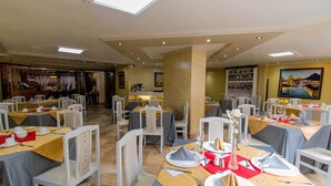 Ein Restaurant mit mehreren gedeckten Tischen, ausgestattet mit einer Mischung aus weißen und beige farbenen Stühlen sowie unterschiedlichen Tischgestaltungen mit Tellern, Besteck und Gläsern.