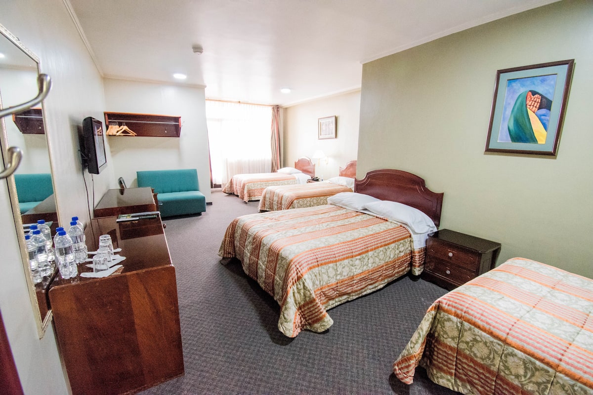Habitación de hotel con tres camas, un escritorio con botellas de agua, un espejo y un cuadro en la pared.