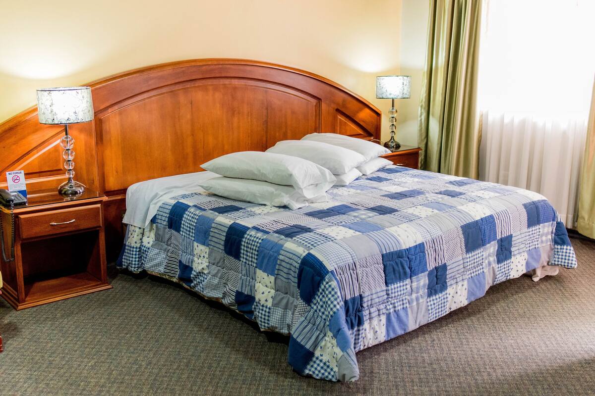 Habitación de hotel con cabecera de madera, colcha a cuadros, dos mesitas de noche con lámparas y una ventana con cortinas.