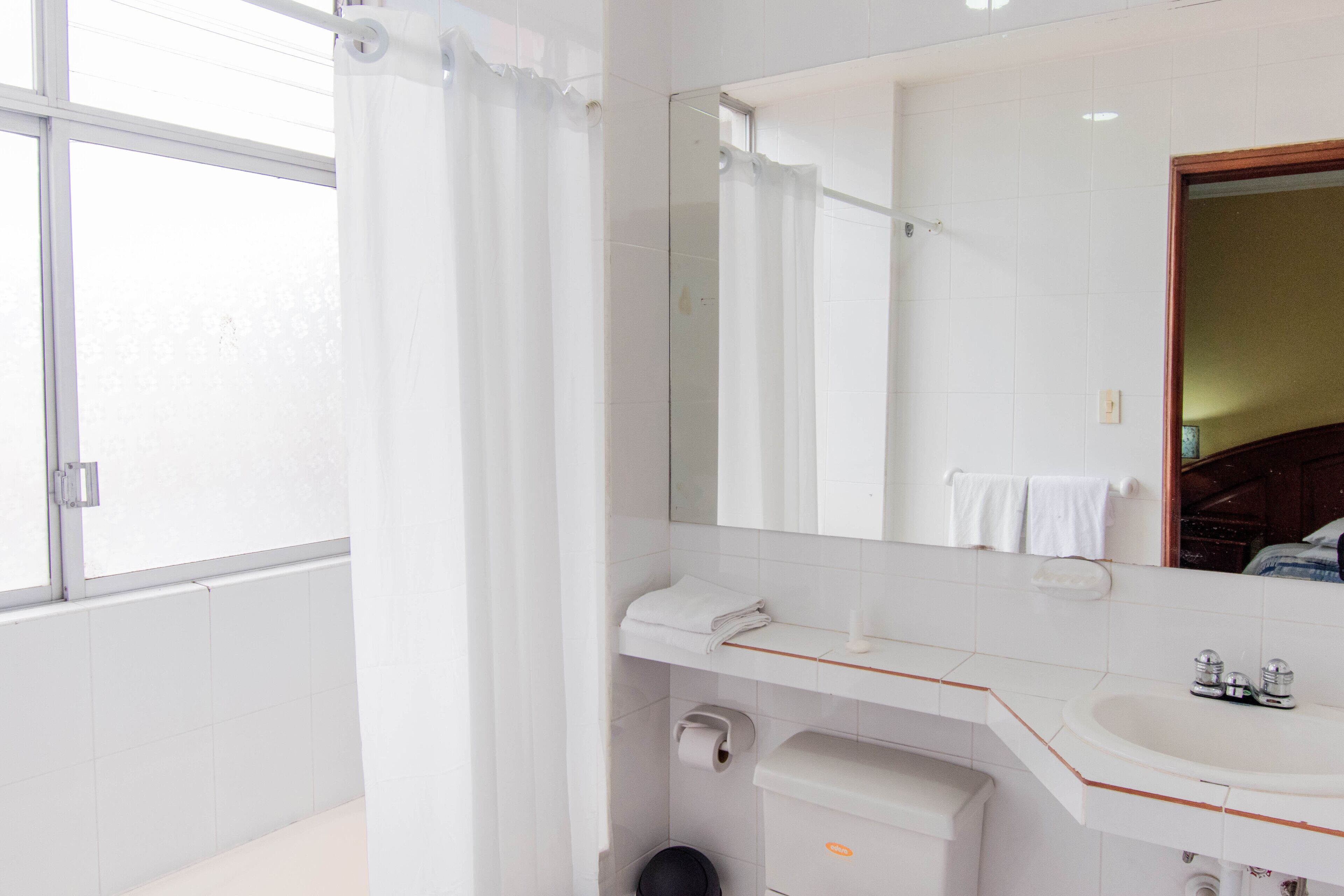 A modern bathroom with a large mirror, white sink, and a window with a curtain.