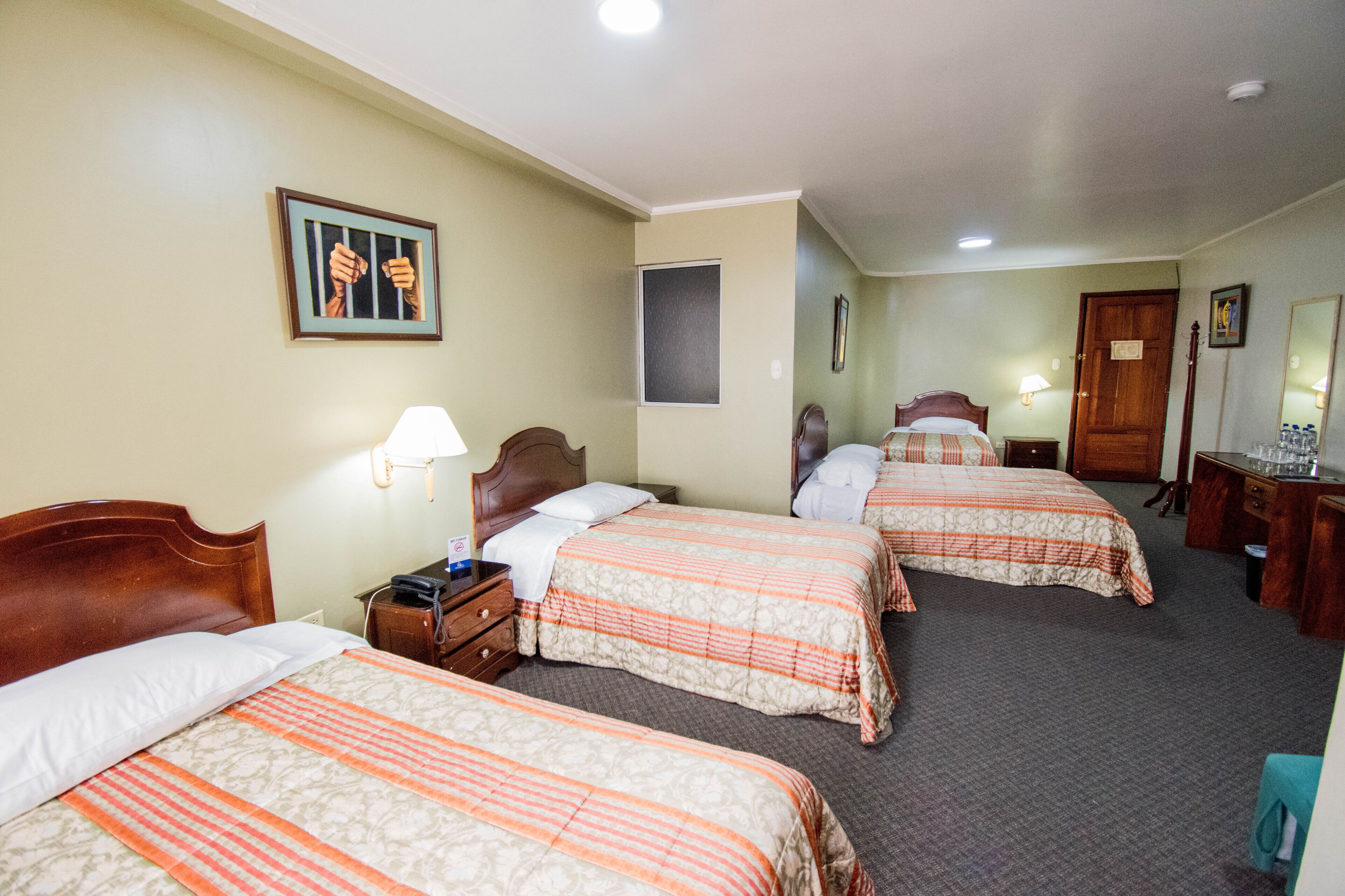 Une chambre d’hôtel avec trois lits, une tête de lit en bois, une table de chevet avec une lampe, un bureau avec un miroir et un tableau encadré au mur.