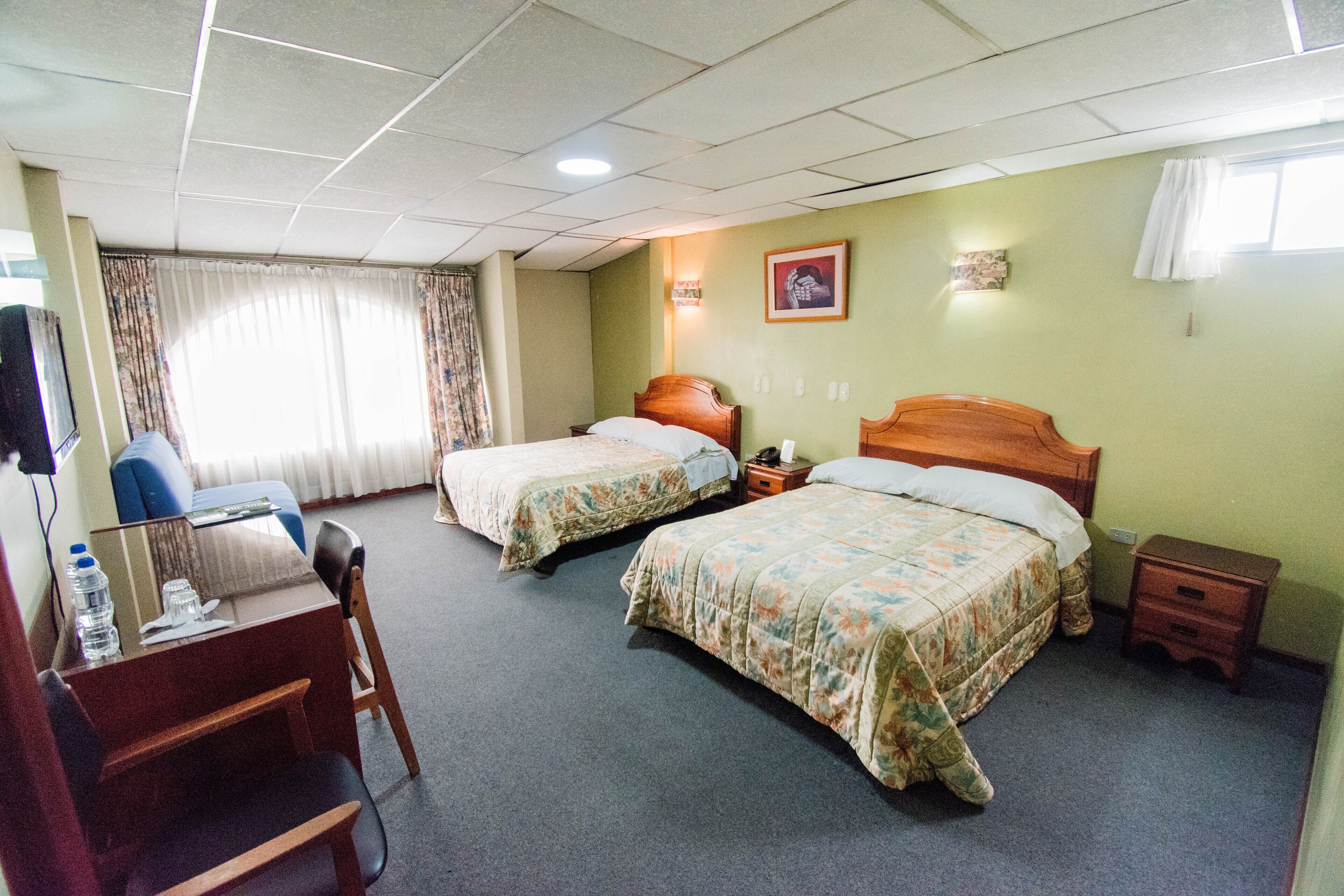 A hotel room with two beds, a desk with a chair, a small table, and a window with curtains.