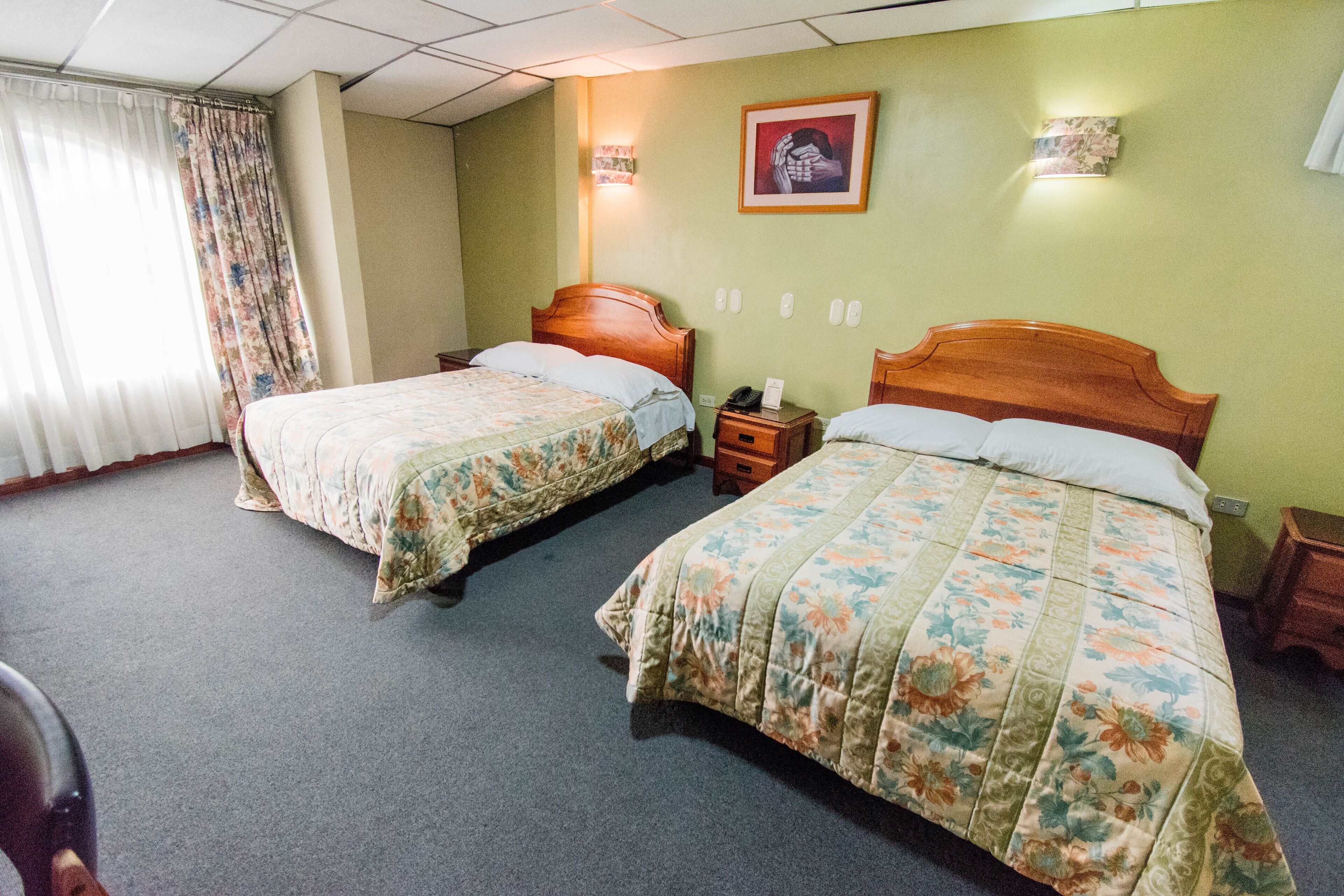 Habitación de hotel con dos camas, cabecero de madera, mesita de noche y lámpara de pared.
