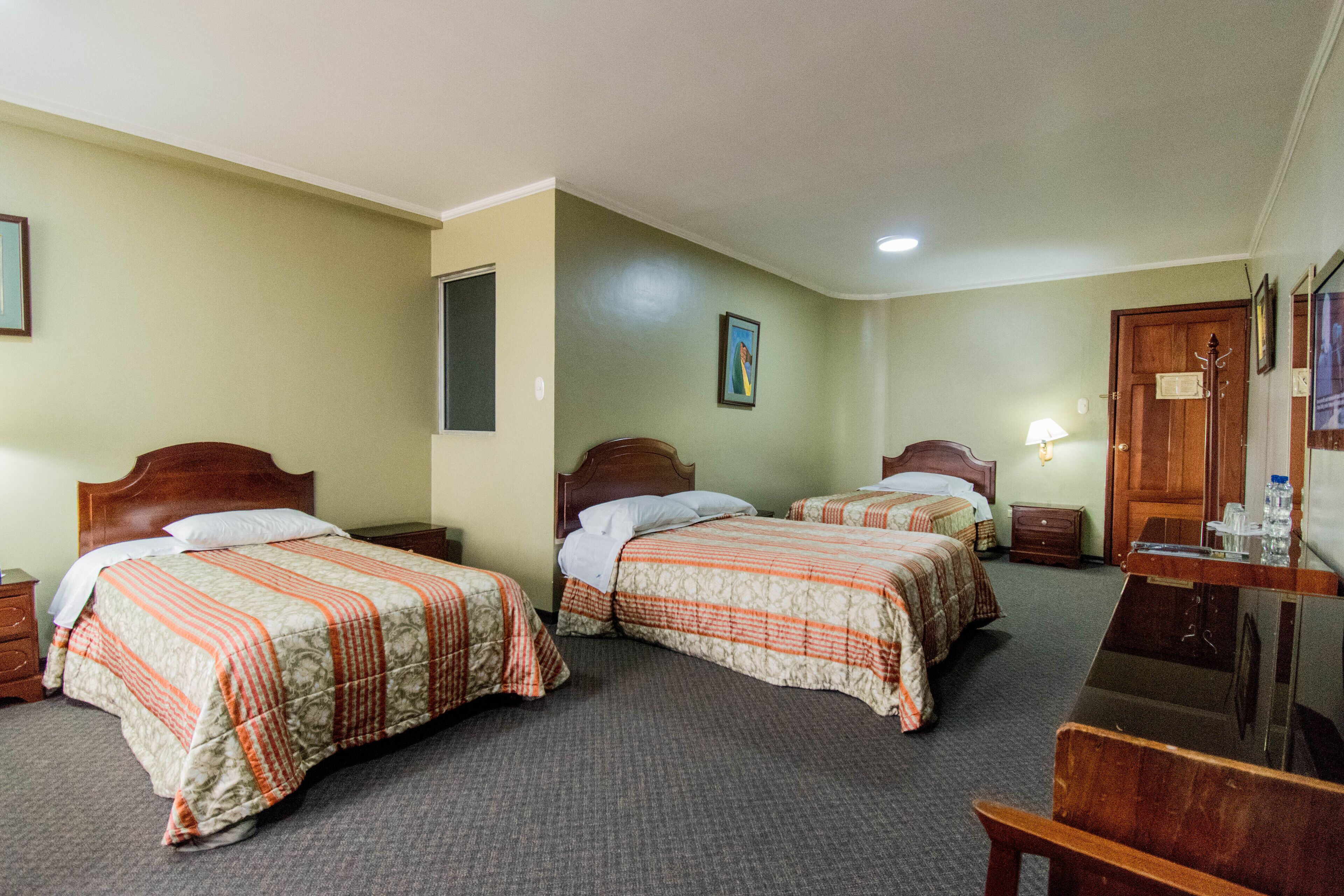 Une chambre d’hôtel avec deux lits simples, un bureau en bois, une chaise, une table de chevet et une lampe fixée au mur.