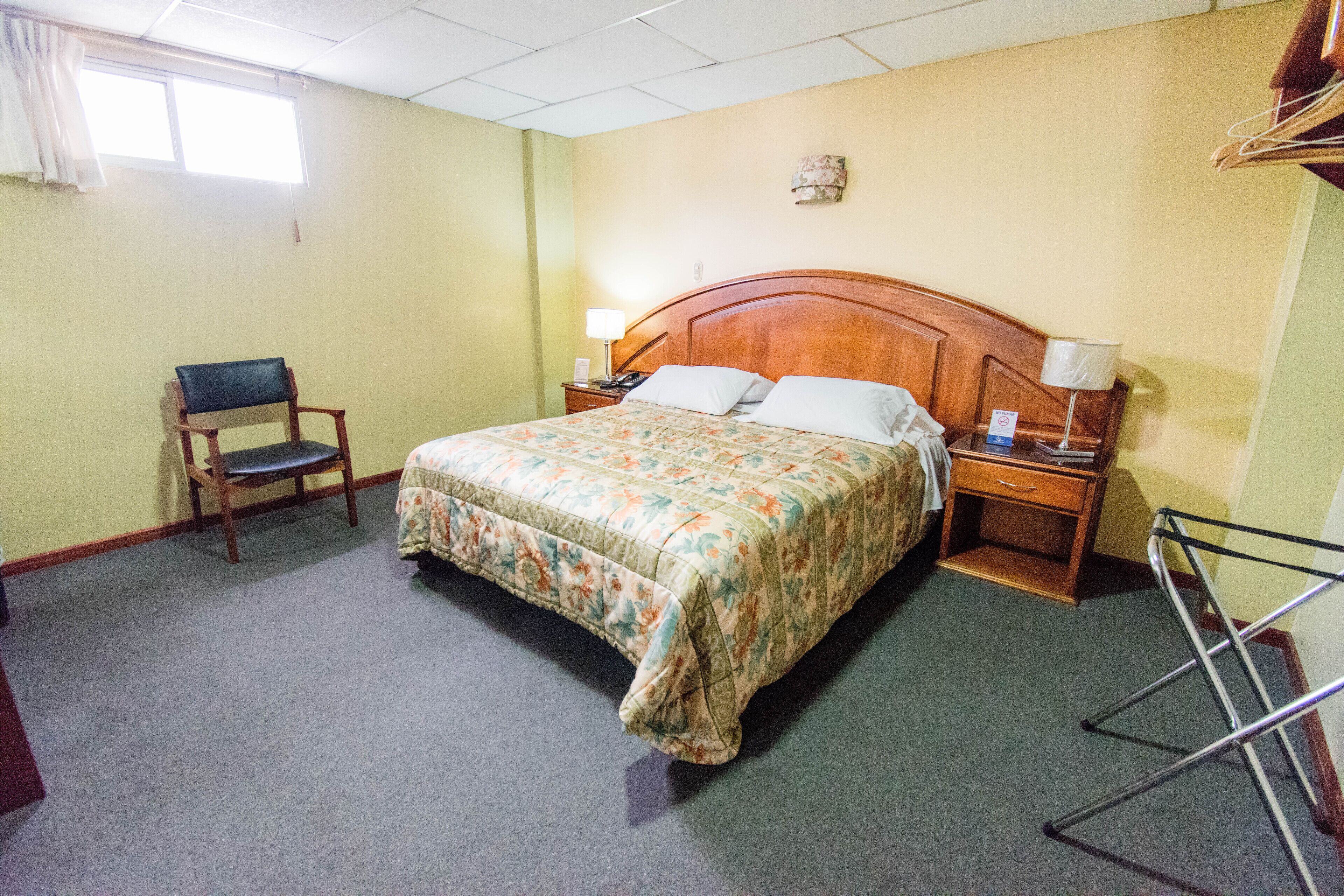 A hotel room with a bed, a chair, a nightstand, a lamp, and a window.