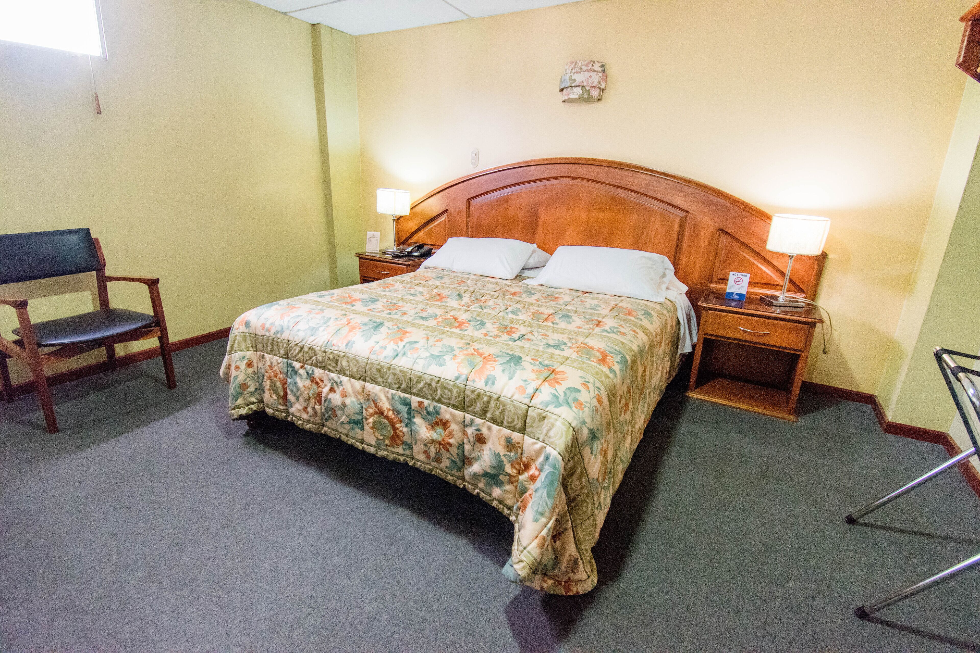 A hotel room with a wooden headboard, a floral bedspread, a nightstand with a lamp, and a chair.