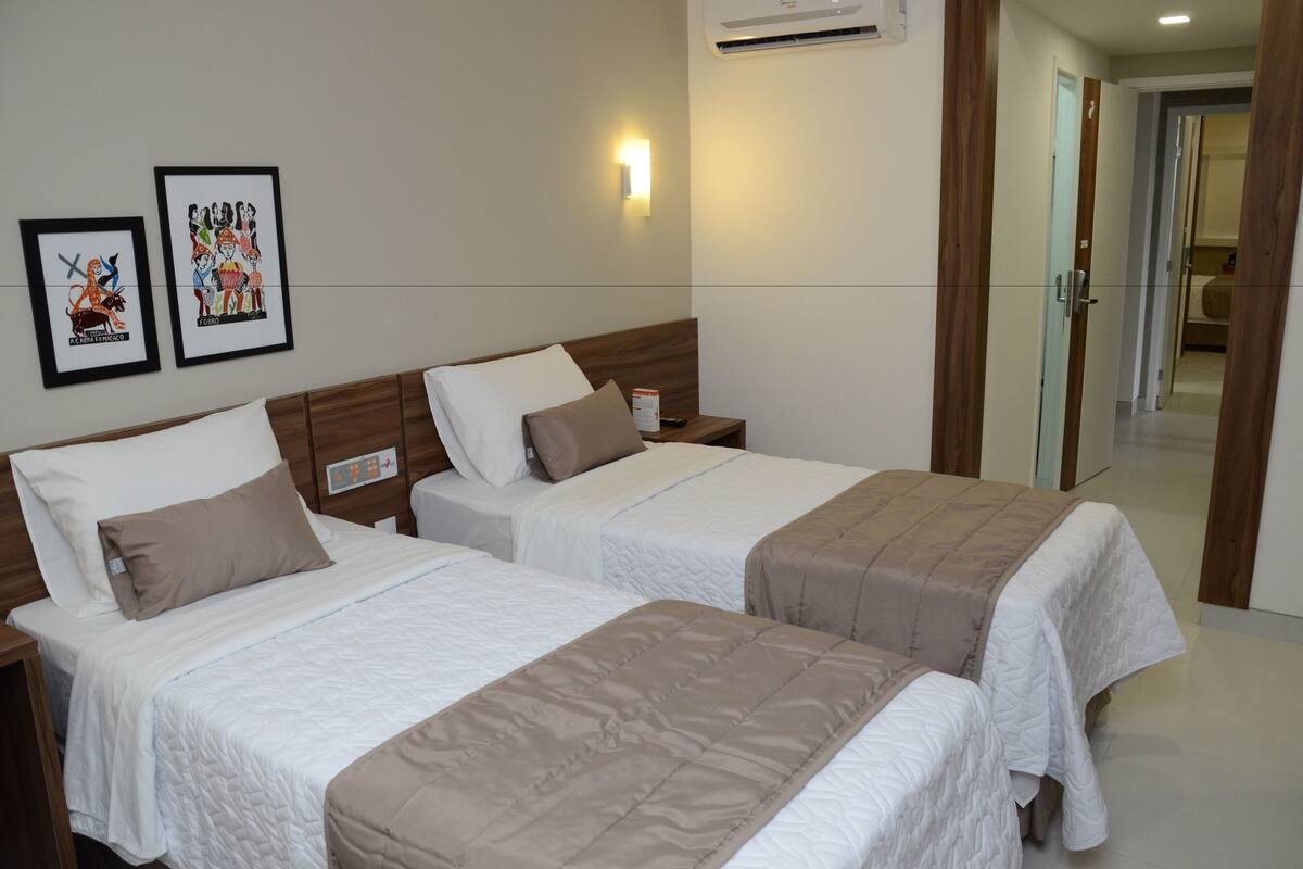 Two single beds with wooden headboards, white bedding, and a bedside table with a lamp. There are framed pictures on the wall and a door leading to another room.