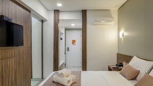 Habitación de hotel con cama, televisor montado en la pared, mesita de noche con teléfono y control remoto, y baño visible a través de una puerta de vidrio.