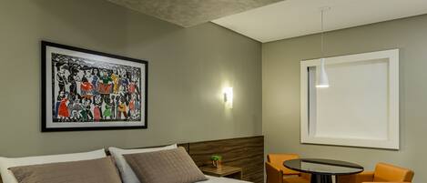 A hotel room with a bed, a framed artwork, a small table with a chair, and a window.