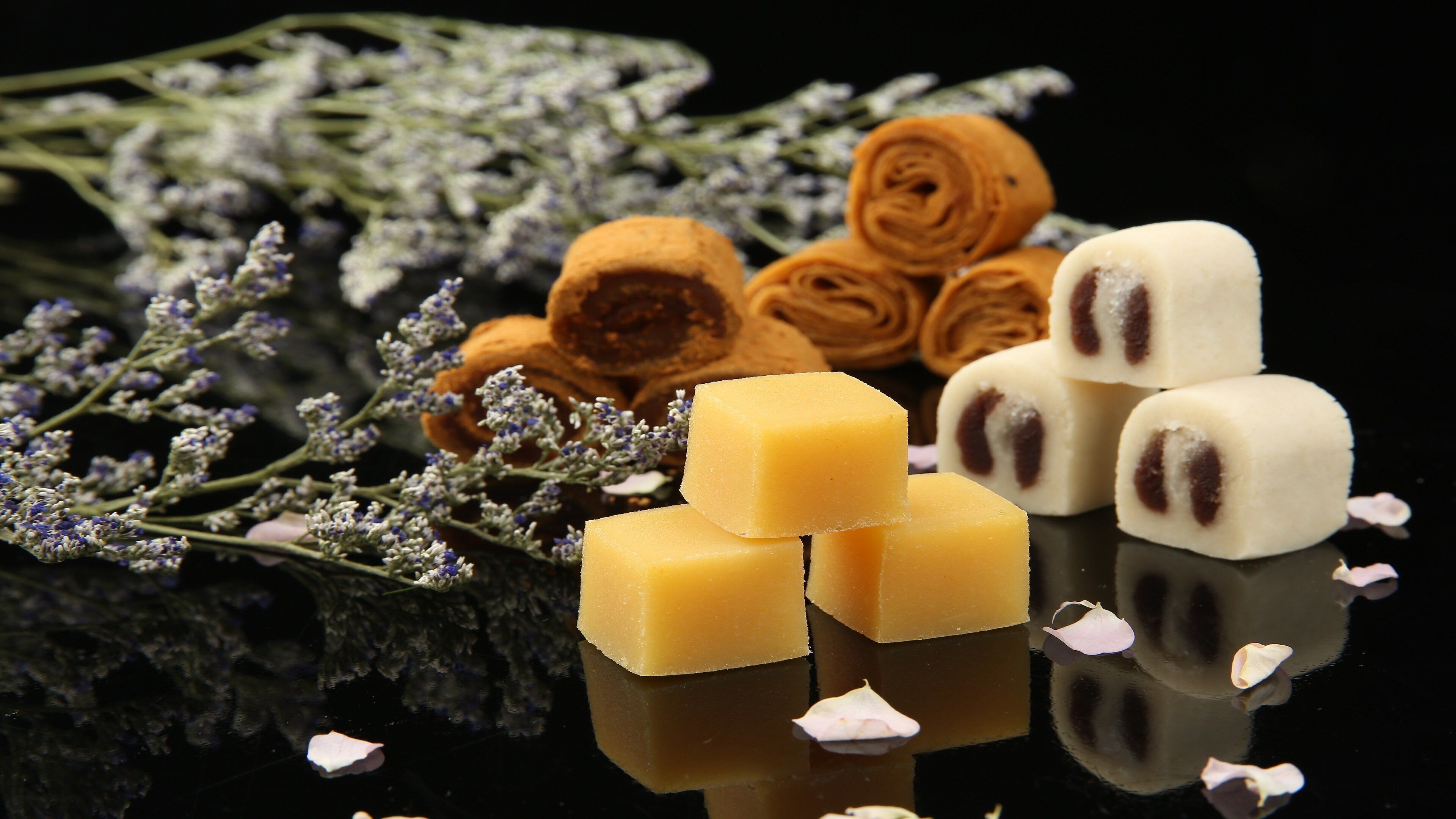 A close-up of food items, including cubes of cheese and chocolate-covered treats, with lavender flowers and rolled pastries in the background.