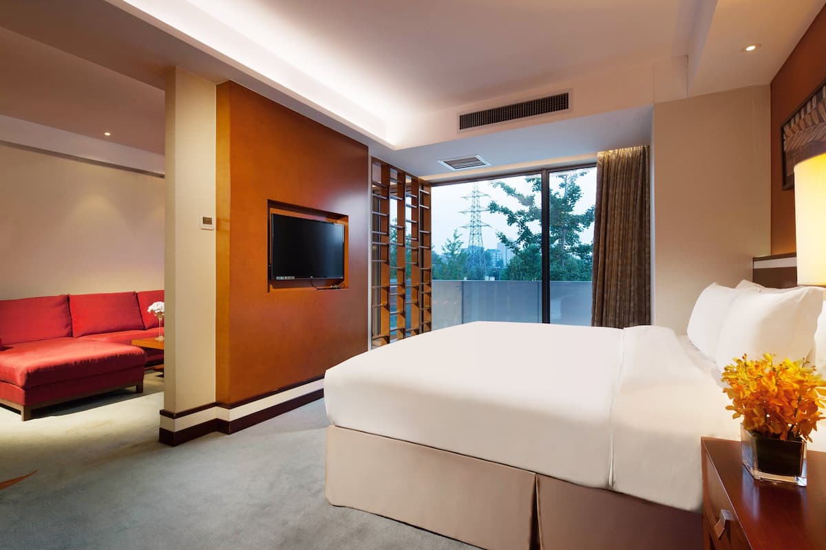 A hotel room with a large bed, a red sofa, a TV, and a window with a view of trees and power lines.