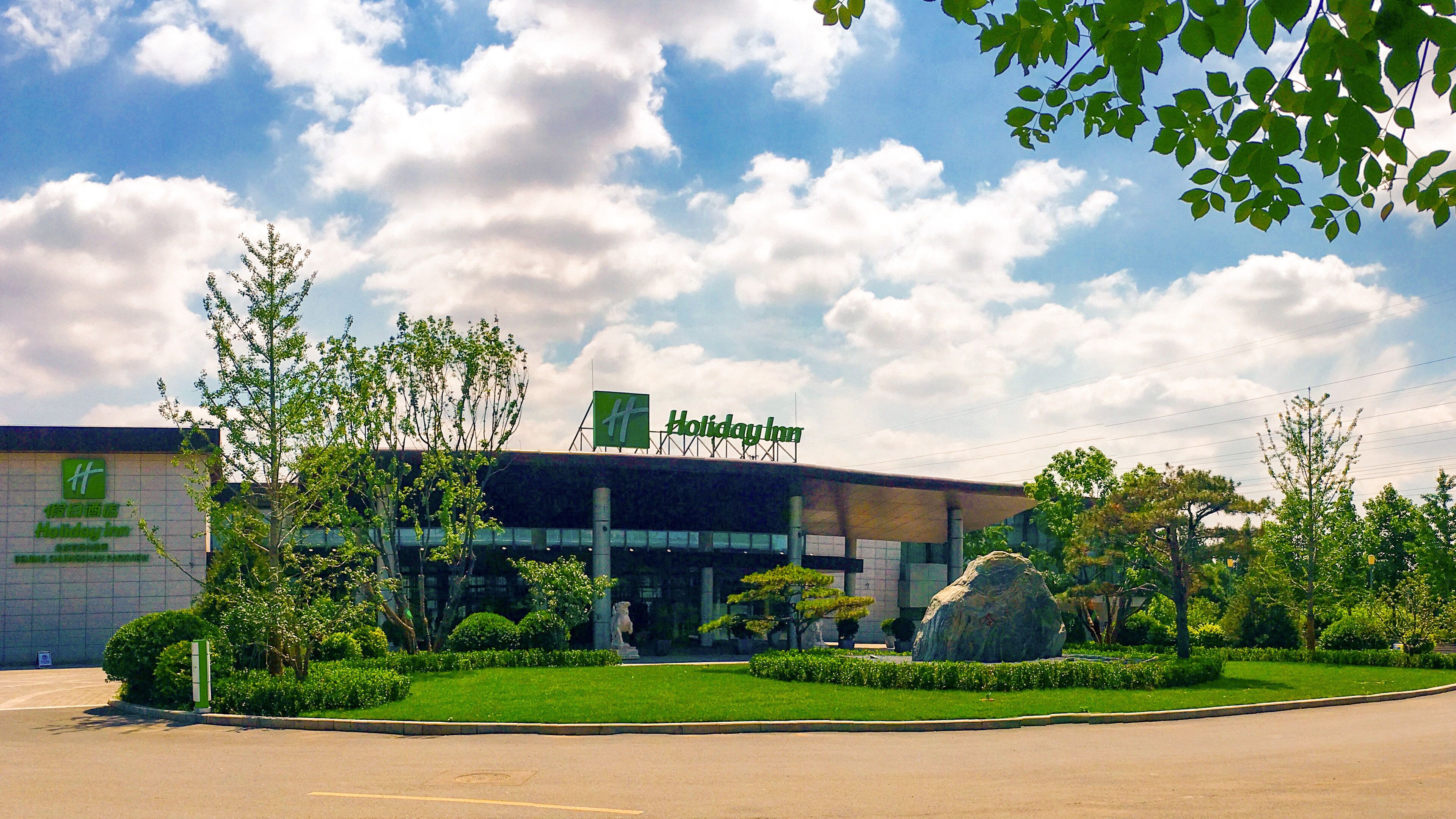 A Holiday Inn hotel with a modern facade, a large rock feature in the front garden, and a clear sky with clouds.