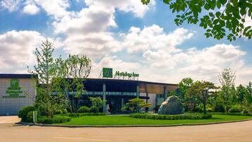 A Holiday Inn hotel with a modern facade, a large rock feature in the front garden, and a clear sky with clouds.
