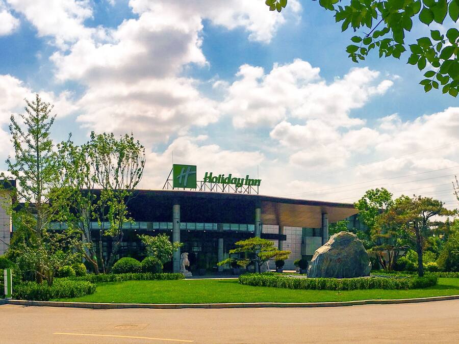 A Holiday Inn hotel with a modern facade, a large rock feature in the front garden, and a clear sky with clouds.