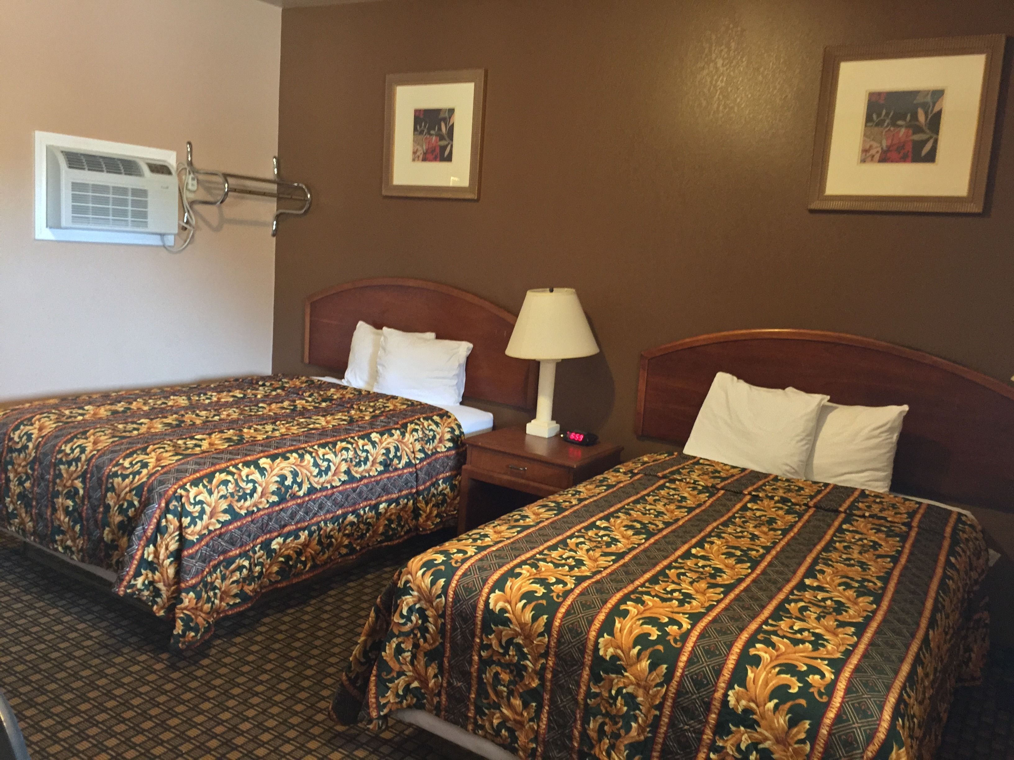 A hotel room with two beds, a nightstand with a lamp, and an air conditioning unit on the wall.