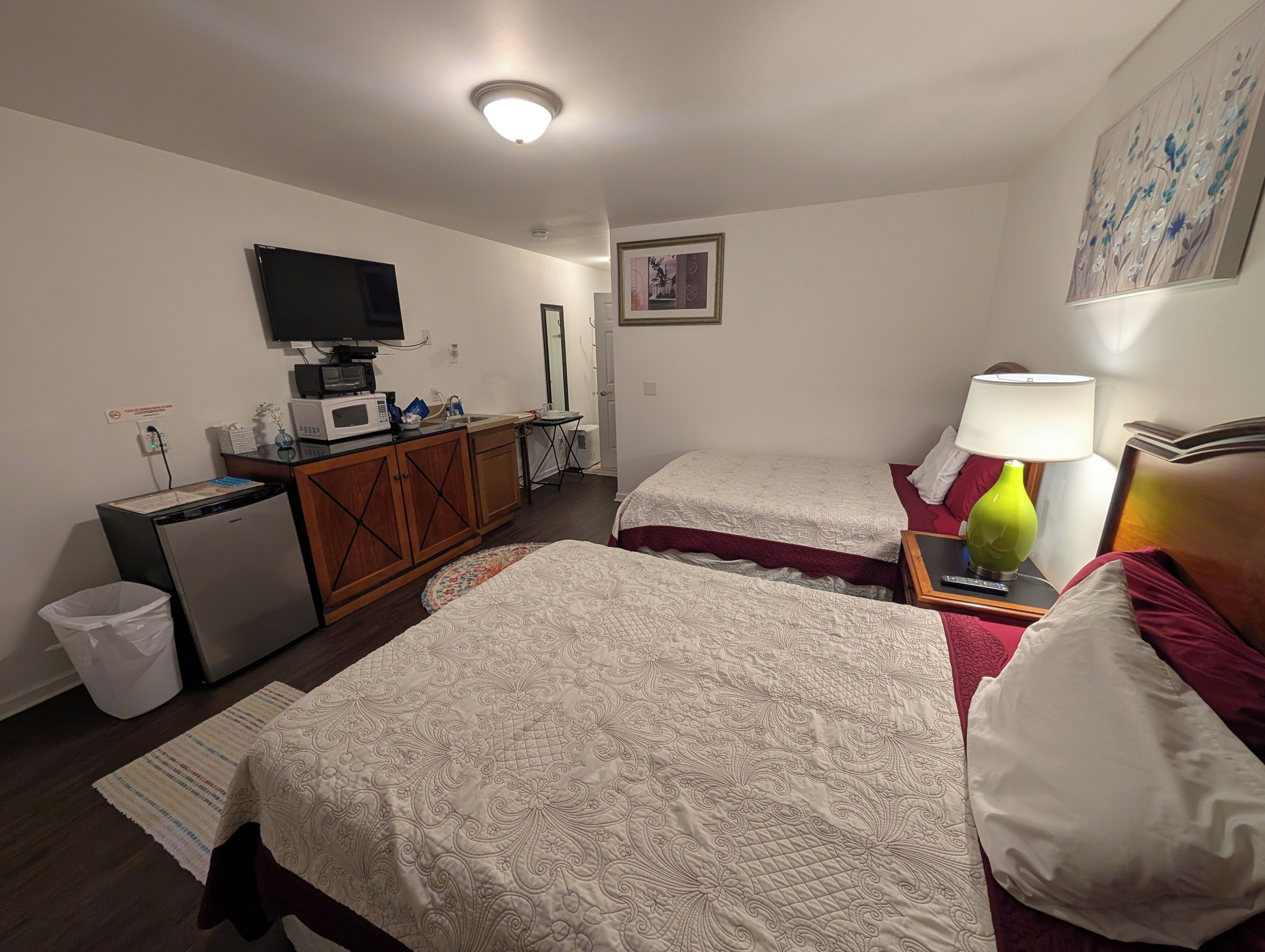 A hotel room with two beds, a small kitchenette, a television, and a painting on the wall.