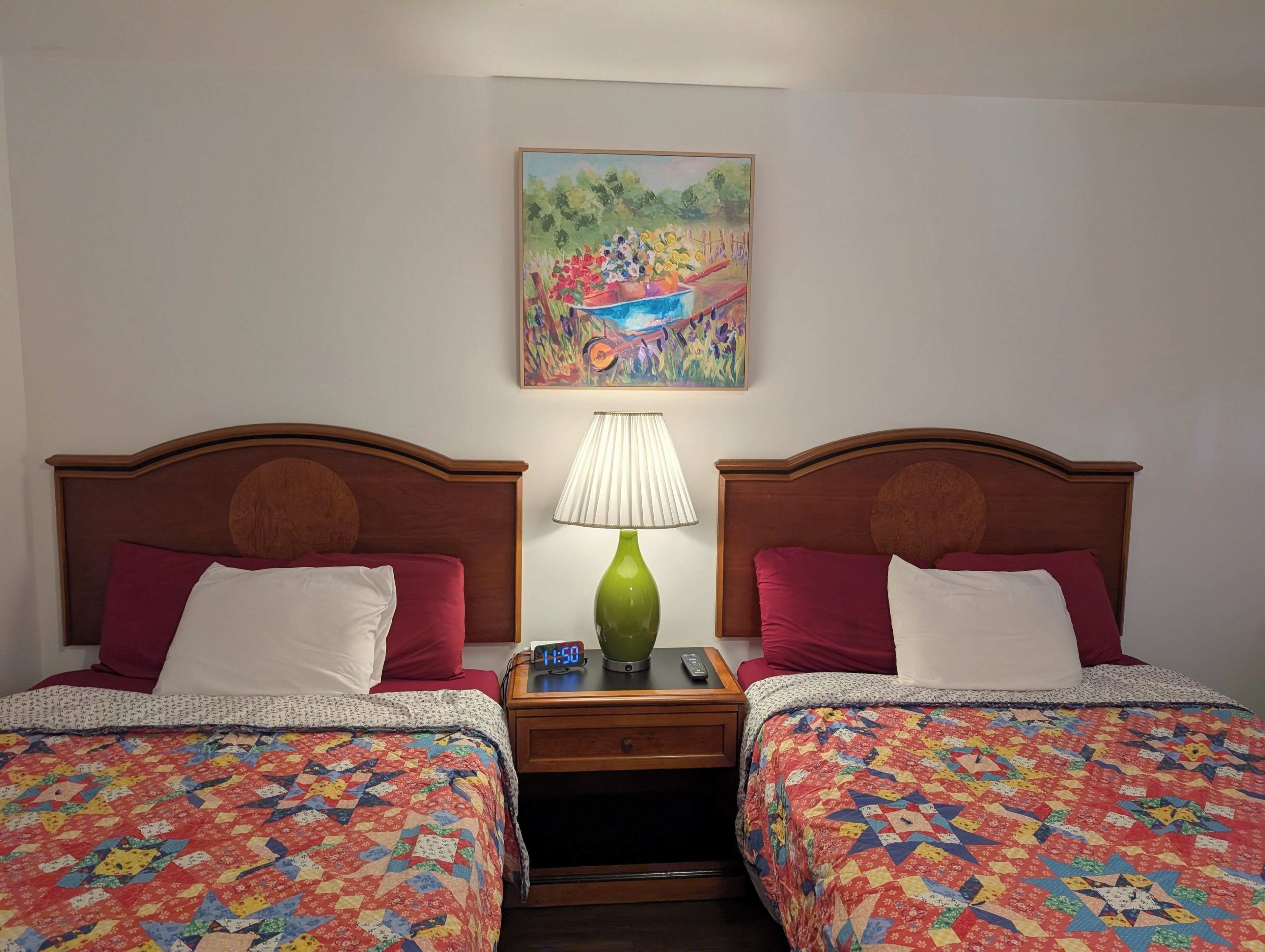 A hotel room with two beds, each with a colorful quilt, a nightstand with a lamp, a clock, and a painting on the wall.