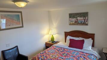 A bedroom with a bed, a chair, a nightstand with a lamp, and two paintings on the wall.