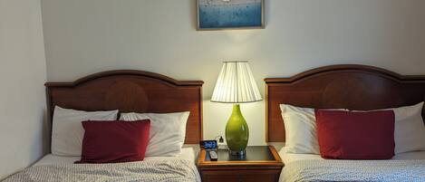 Een hotelkamer met twee bedden, elk met een houten hoofdbord, witte en rode kussens en kleurrijke dekens. Daartussen staat een nachtkastje met een groene lamp en een digitale klok. Boven de bedden hangt een schilderij.