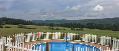 A swimming pool surrounded by a white picket fence, with a view of green fields and hills in the background.