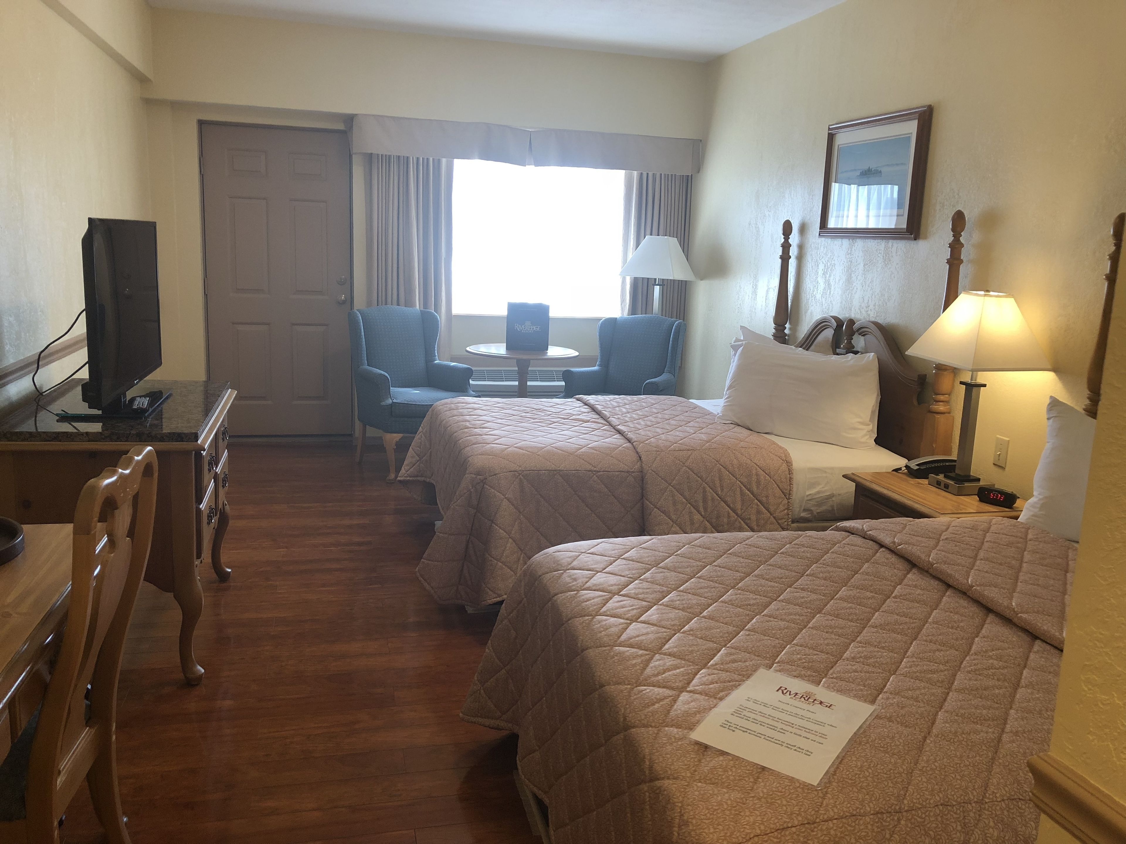 A hotel room with two beds, a desk with a television, a chair, a table, and a window with curtains.