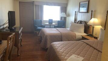 Quarto de hotel com duas camas, uma escrivaninha com televisão, uma cadeira, uma mesa e uma janela com cortinas.