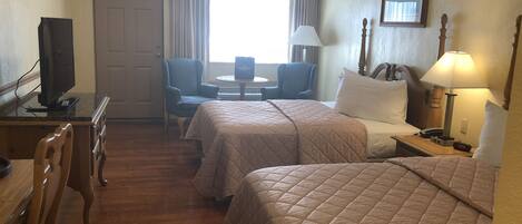 A hotel room with two beds, a desk with a television, a chair, a table, and a window with curtains.