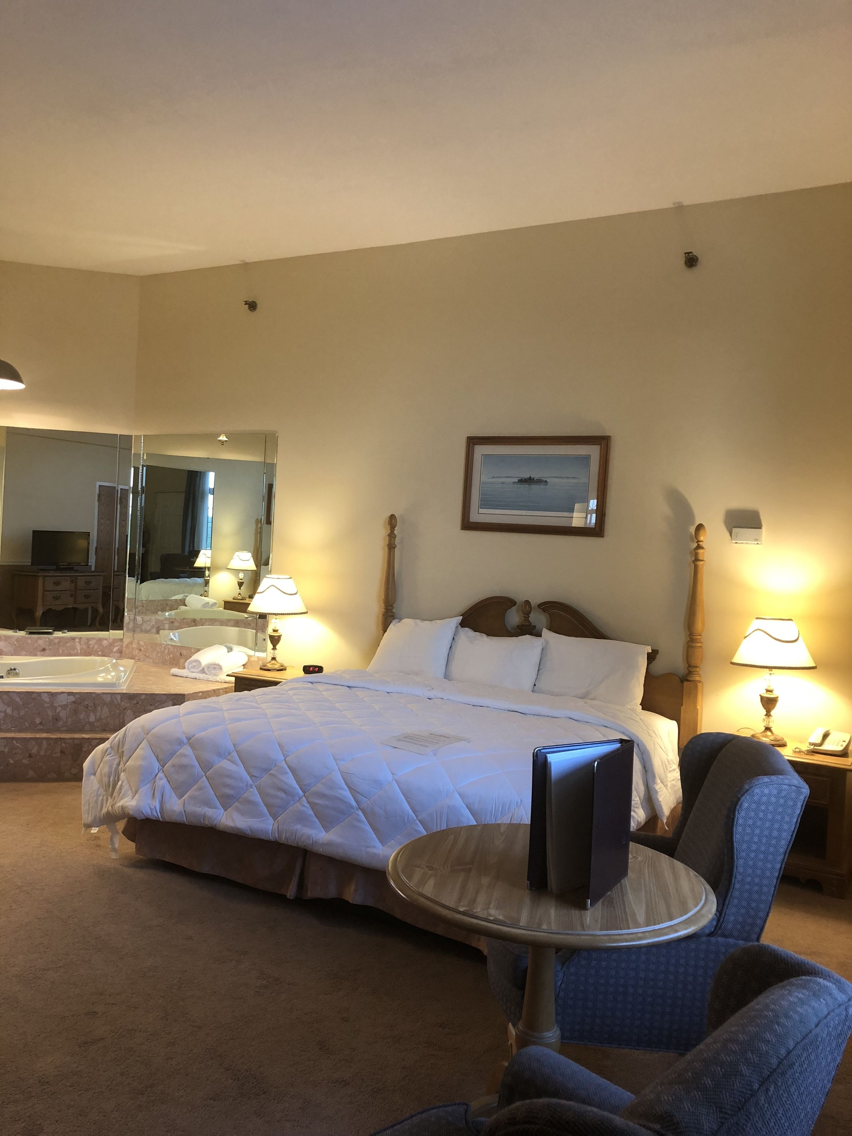 Una camera d'albergo con un letto, una sedia, un tavolino, uno specchio, un quadro appeso al muro e un bagno visibile nello specchio.