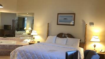 Habitación de hotel con cama, una silla, una mesita, un espejo, un cuadro en la pared y un baño visible a través del espejo.