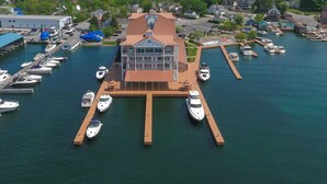 A marina with multiple boats docked, a large building with a distinctive roof, and a backdrop of residential houses and greenery. - Riveredge Resort (Alexandria Bay)