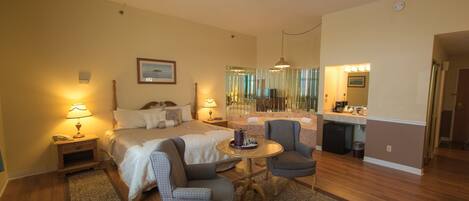 A hotel room with a bed, two chairs, a small table, a desk with a mirror, and a kitchenette.