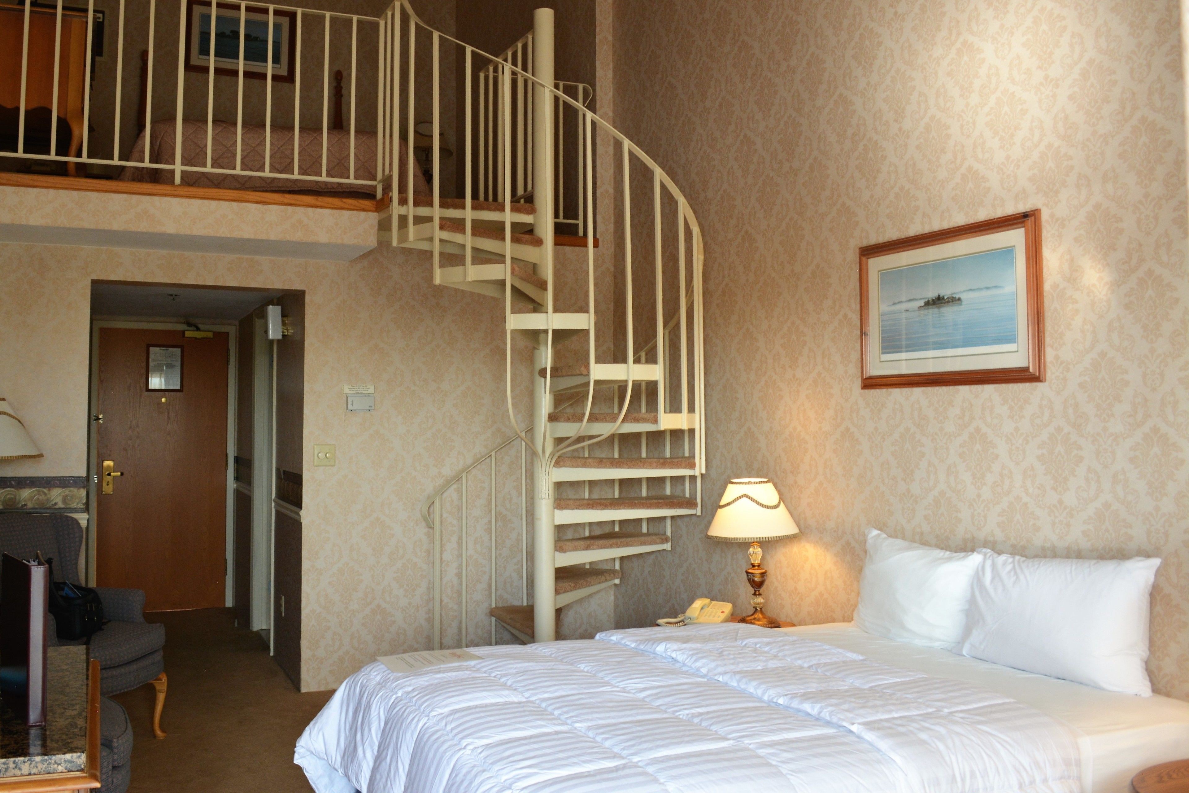 A hotel room with a bed, a desk, a chair, a desk lamp, a telephone, a picture frame, and a spiral staircase.