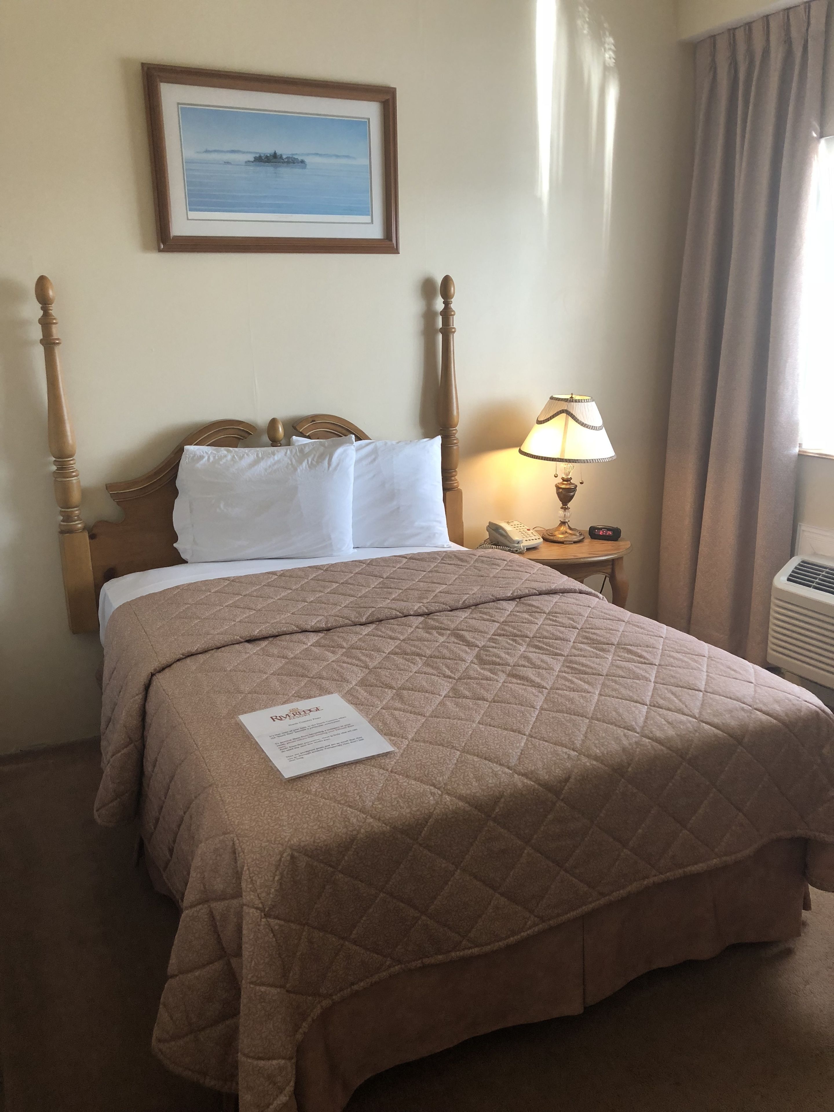 Uma cama bem arrumada com um edredom marrom, um quadro na parede, uma luminária de cabeceira e um telefone sobre uma mesa pequena ao lado da cama.