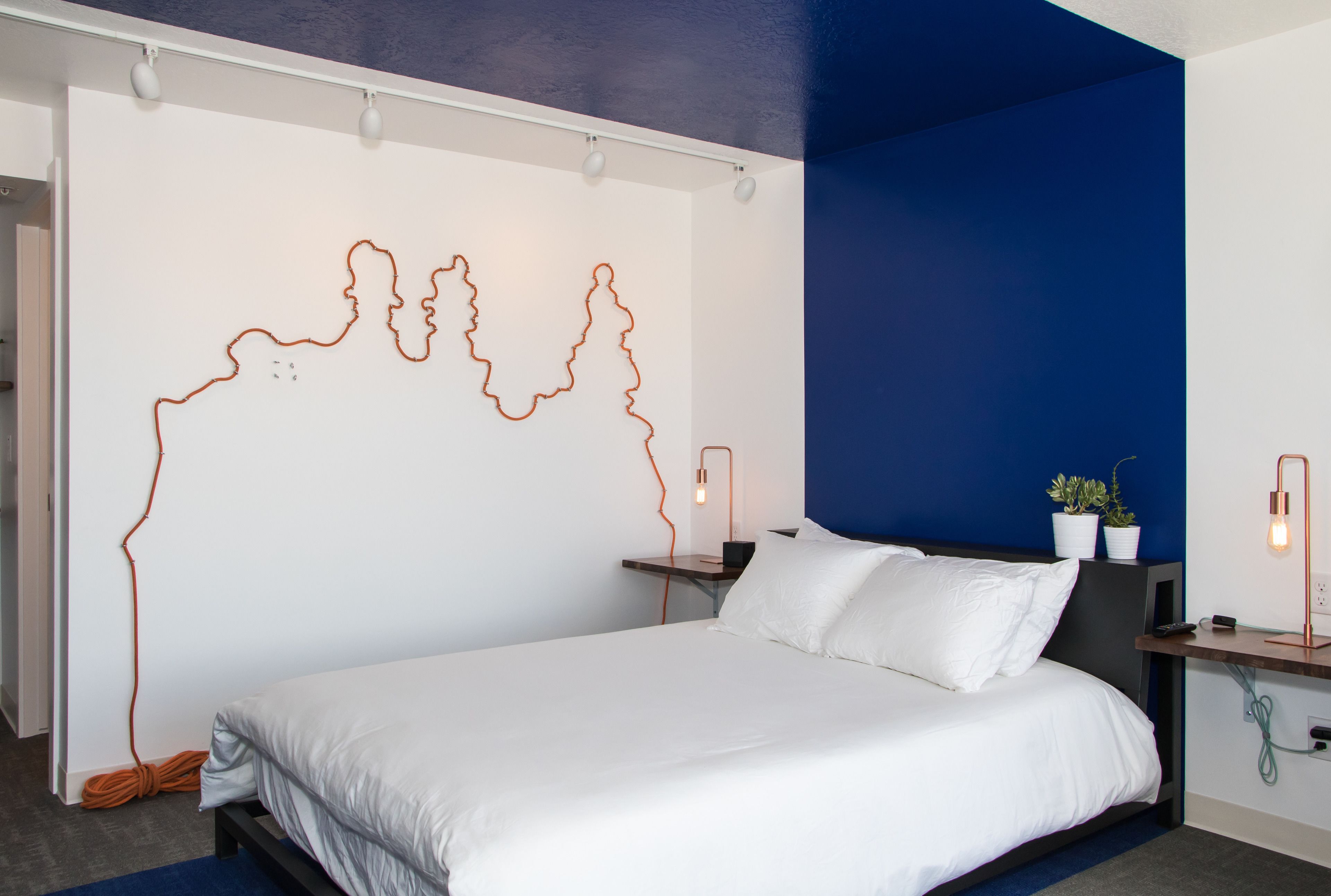 A modern hotel room with a large bed, a wall art installation, bedside tables with lamps, and a blue accent wall.