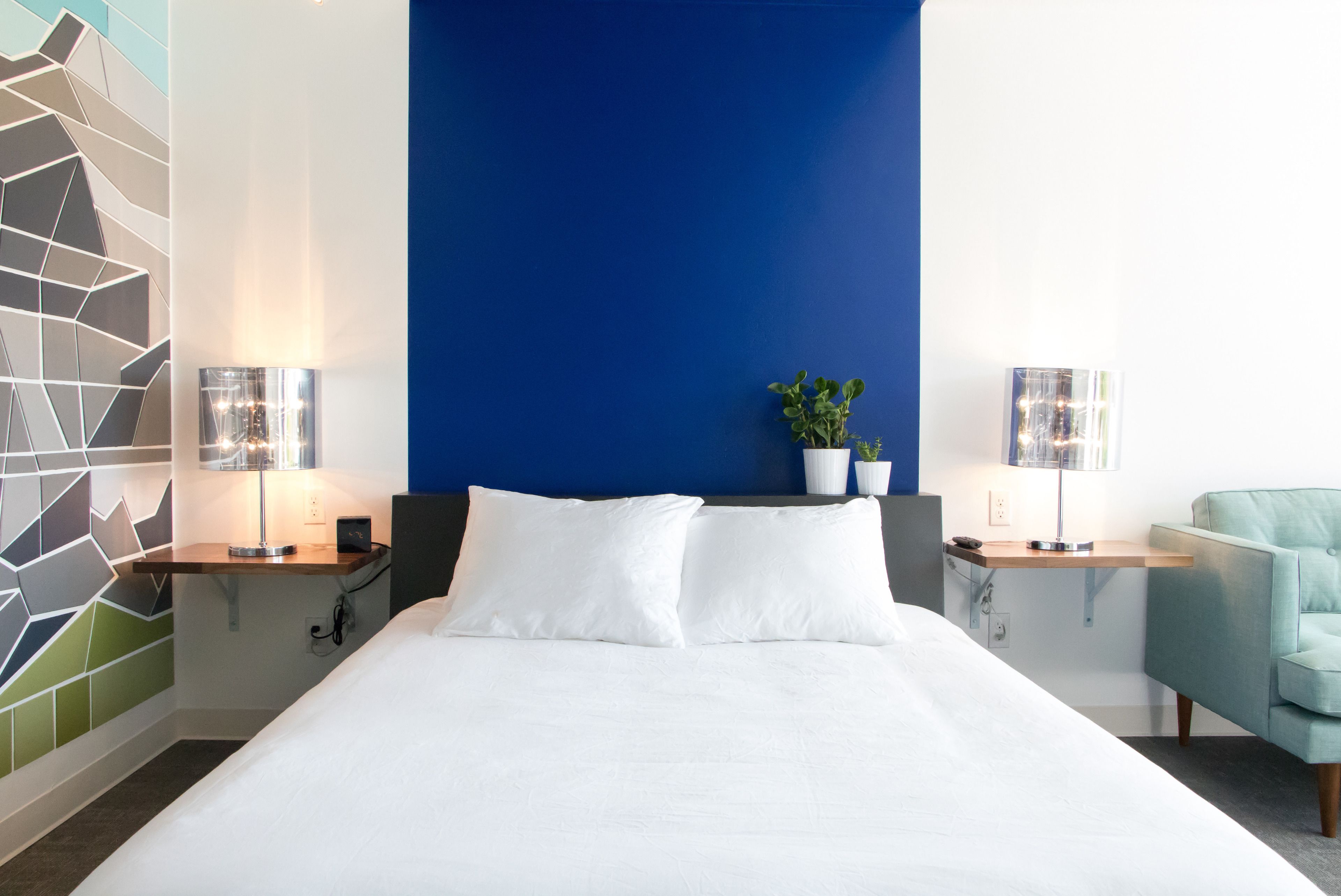 A modern hotel room with a large bed, two bedside tables with lamps, a blue accent wall, and a teal armchair.