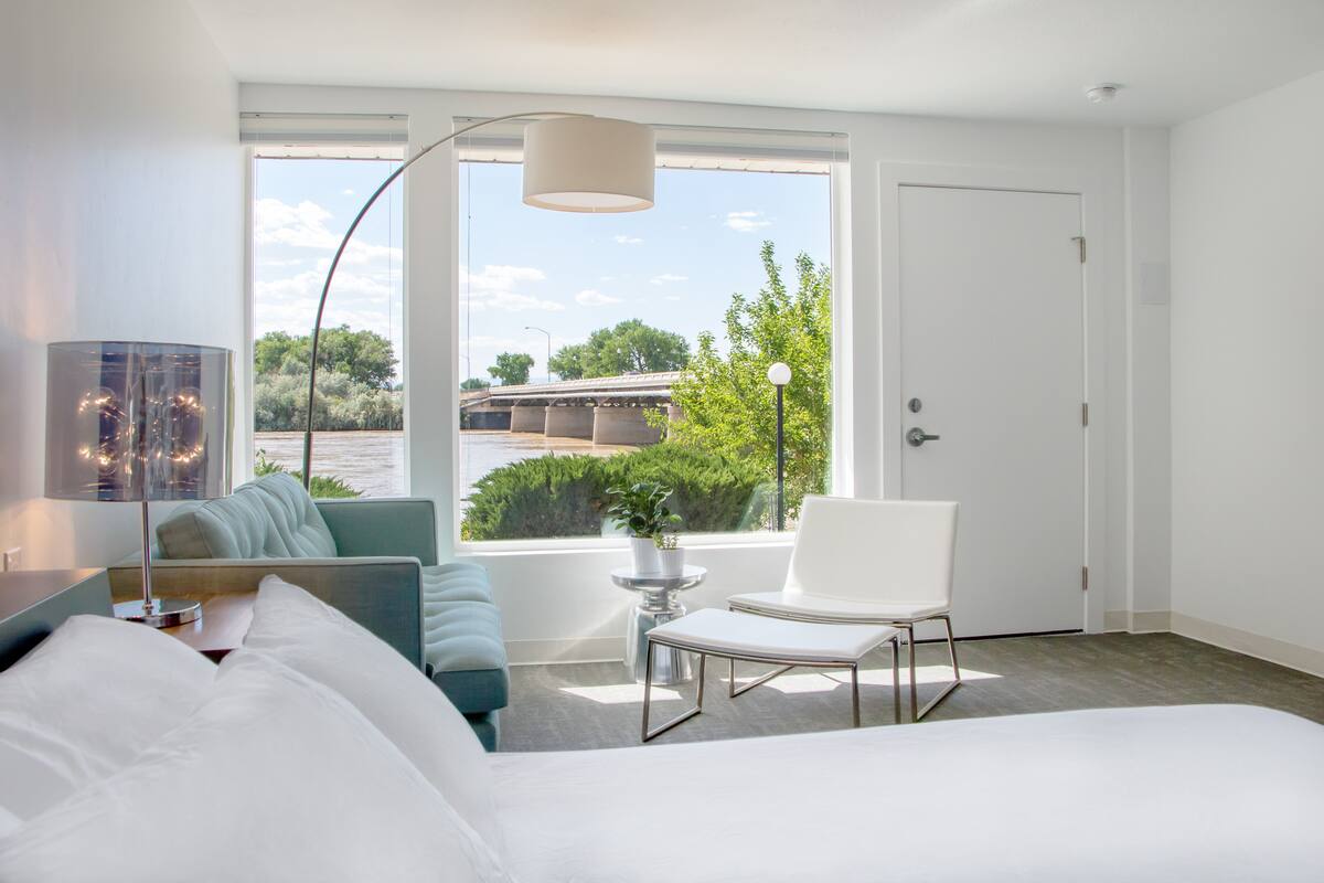 A modern hotel room with a bed, a sofa, a chair, a lamp, and a view of a bridge and greenery.