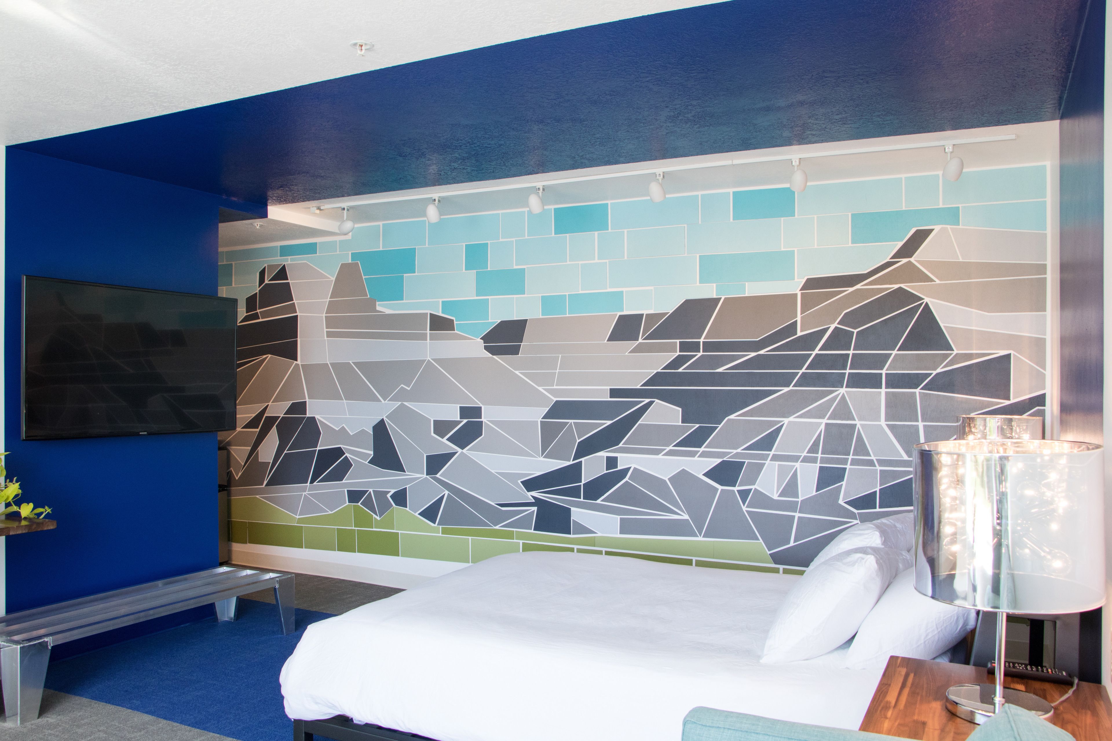 A hotel room with a bed, a bench, a TV, and a wall with a geometric pattern.