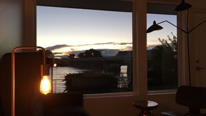 Una habitación con un gran ventanal que da a un río y un puente al atardecer. Hay un escritorio con una lámpara y una silla.