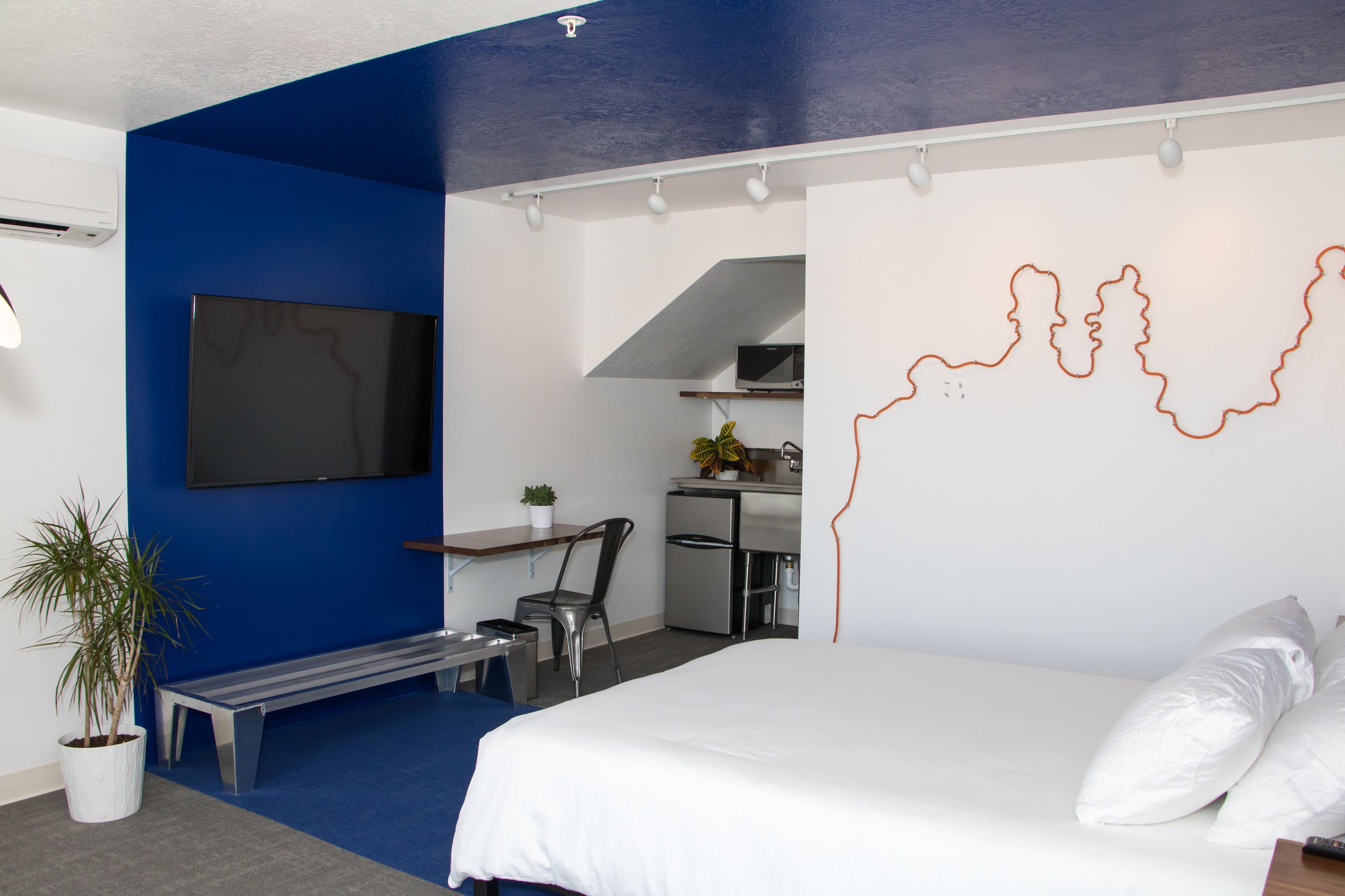 A modern hotel room with a large bed, a desk with a chair, a small kitchenette, and a TV mounted on the wall.