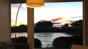 Una habitación con un ventanal grande que da a un río al atardecer, un sofá y una lámpara.