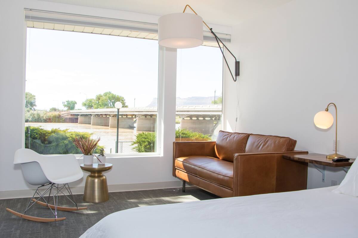 A modern hotel room with a large window, a brown leather sofa, a white chair, a small round table with a plant, and a bedside lamp.