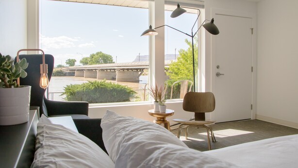Una habitación de hotel moderna con un ventanal grande, una cama, un escritorio con silla, una mesita con una planta y una lámpara.