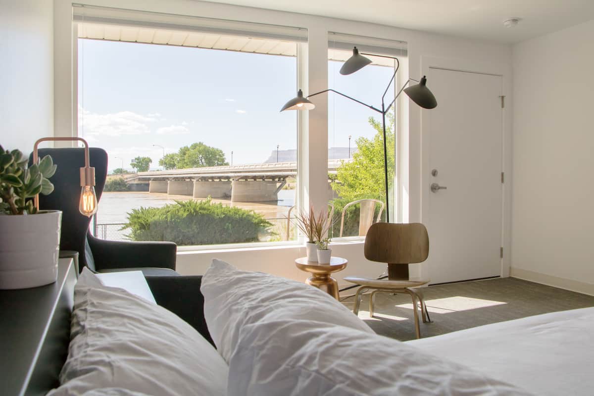 A modern hotel room with a large window, a bed, a desk with a chair, a small table with a plant, and a lamp.