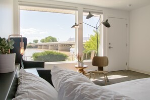 A modern hotel room with a large window, a bed, a desk with a chair, a small table with a plant, and a lamp. - Skyfall Guestrooms (Green River)