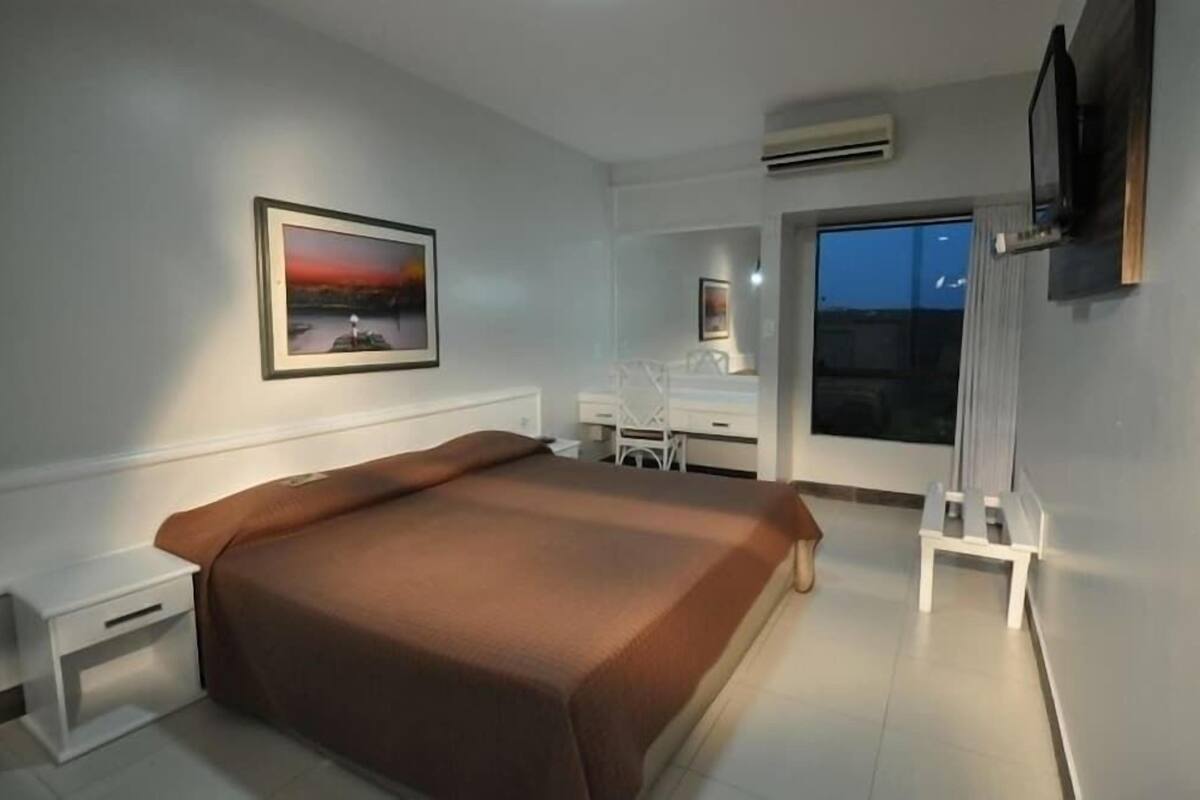 A bedroom with a bed, a desk, a chair, a TV, and a window.