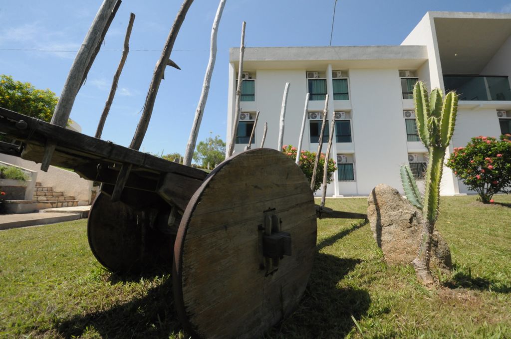 A large wooden barrel with a metal mechanism, a cactus, and a building with multiple windows and air conditioning units in the background.